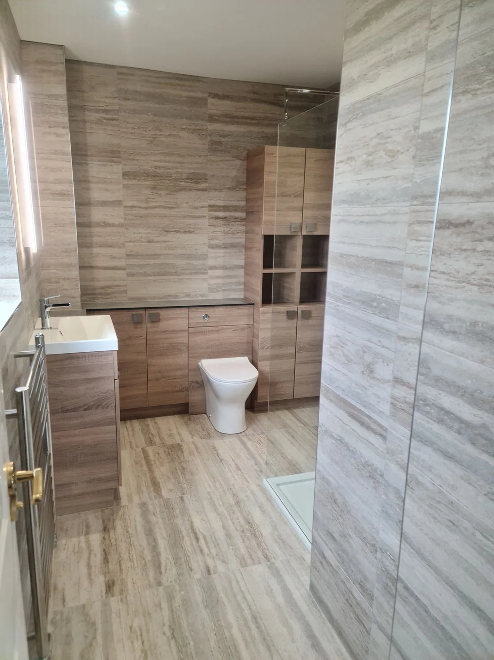Bathroom tiled in warm travertine with fitted wood-effect vanity units and a walk-in shower
